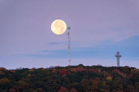 A supermoon rises above autumn treetops and a cross, with a radio tower silhouette. g.の写真素材