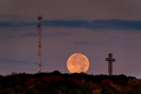 A supermoon rises above autumn treetops and a cross, with a radio tower silhouette. g.の写真素材