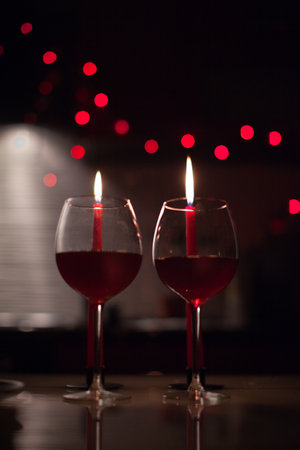 Two red wine glasses with lit candles behind, warm bokeh lights and romantic mood. g.