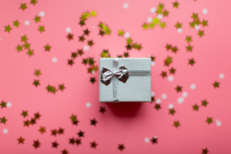 Silver gift box with a glitter ribbon on a pink festive background with golden star confetti.