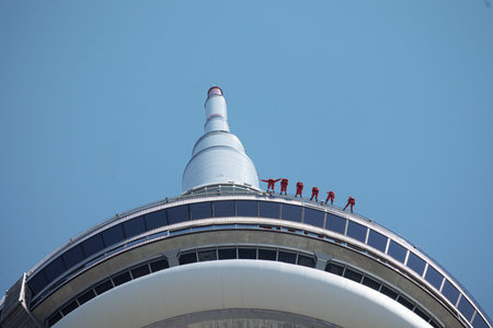 Tourists in red suits on EdgeWalk attraction at CN Tower observation deck against blue sky in...