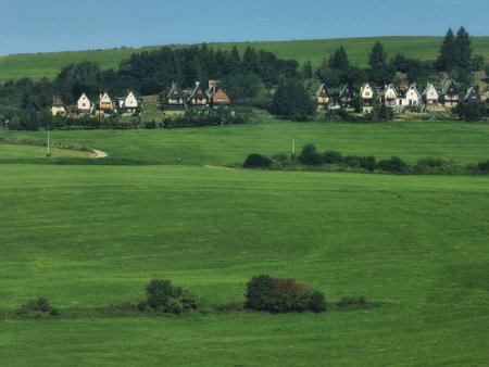 Scenic rural landscape with wide grassy fields, dirt road, and small village houses surrounded by trees under a clear blue sky.の写真素材
