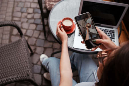 Young Brunette Woman Making Photo of Coffee on Smartphone While Sitting in Cafe Outdoors