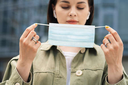 Young Woman Putting a Protective Face Mask on Her Face Because of Pandemic, Portrait of Brunette Girl Using Earphones Standing Outdoors Against City Buildings, Focus on the Maskの写真素材