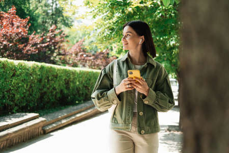 Young Woman Texting on Her Smartphone While Walking in the City Park, Brunette Girl Using Cell Phone...