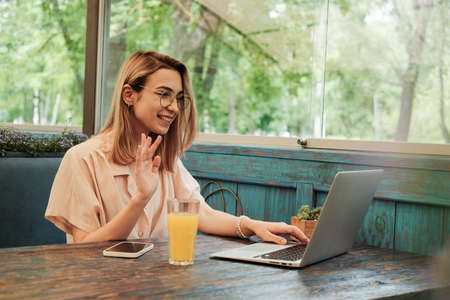 Young Confident Woman Using Laptop for Video Call and Greeting by Hand While Sitting in Restaurant,...