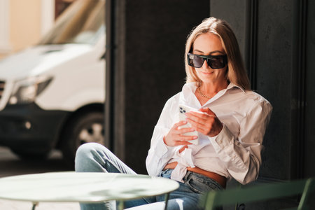 Elegant Caucasian Woman Sitting at a Cafe Table and Texting on Smartphone, Confident Female Entrepreneur Wearing Sunglasses and White Shirt While Waiting for Meetingの写真素材