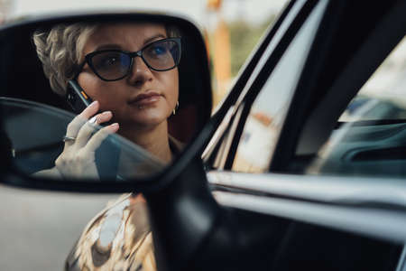 Stylish Woman in Glasses Talking on a Phone While Driving Car, View Through Rear Mirrorの写真素材