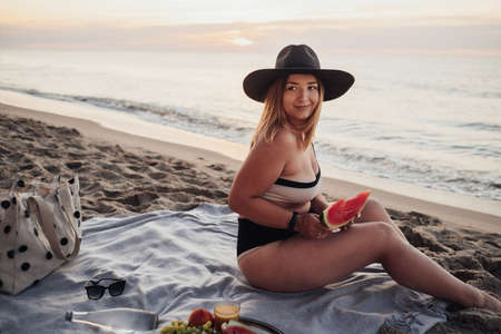Young Pretty Woman Holding Slice of Watermelon and Looking Away While Sitting on Sandy Beach by the Sea at Sunriseの写真素材