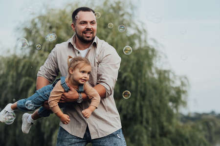Happy Young Father Holding His Daughter in Arms and Imitating Flying and Playing with Bubbles...
