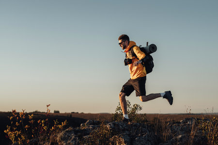 Travel Man Dressed in Windbreaker with Backpack and Camping Mat, Jumping on the Top of Hill During...
