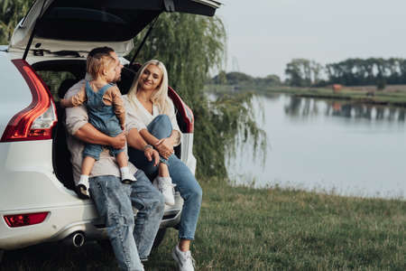 Young Parents with Little Daughter Sitting in Open Trunk of the Car, Happy Family Enjoying Road Trip on Their SUV Carの写真素材