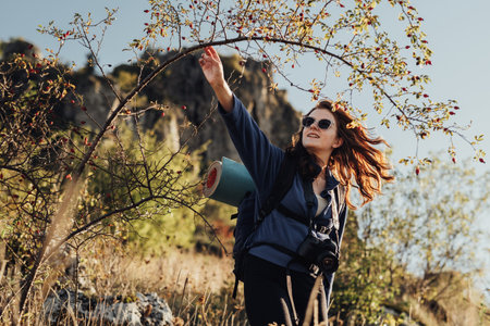 Cheerful Young Travel Woman in Sunglasses With Backpack with Camping Mat and Digital Camera on a Strap Making Her Way Through the Thickets During Her Hikeの写真素材