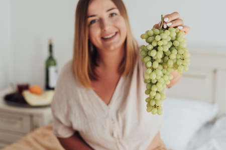 Young Caucasian Woman Holding Bunch of Grapes in Hand While Sitting in Hotel Room, Tray of Fruit and Bottle of Wine Stands on the Backgroundの写真素材
