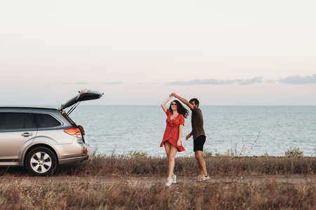 Youth Couple Having Fun on the Background of the Sea, Man and Woman Dancing Near Their SUV Car at Sunsetの写真素材