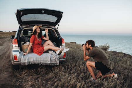 Man Photographing Woman in Red Dress and Hat That Sitting Inside Opened Trunk of the Car on the...