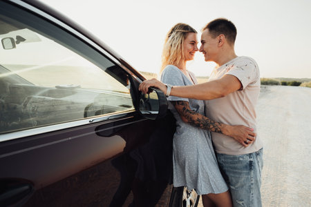 Cheerful Young Couple Hugging Near the Car, Tattooed Woman and Caucasian Man Enjoying Road Trip at Sunsetの写真素材