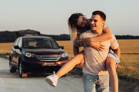 Caucasian Man Carries Girlfriend on His Back on the Background of Their SUV Car, Young Couple Enjoying Their Road Trip at Sunsetの写真素材