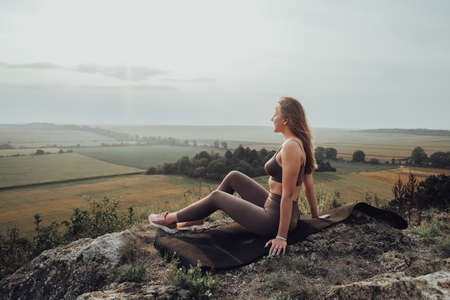 Young Woman Sitting on Top of a Hill Outdoors, Sporty Girl Enjoying Panoramic Landscape at Sunsetの写真素材