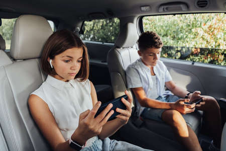 Young Caucasian Girl and Her Teenage Brother Focused on Smartphones While Sitting in Minivan,...