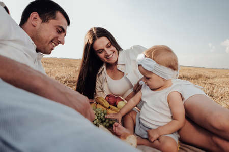 Young Family, Mother and Father with Their Toddler Daughter Having Picnic Time Outdoors in the Field at Sunsetの写真素材