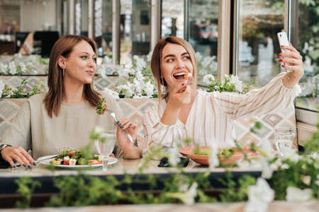 Two Women Making Selfie While Having Lunch Meeting, Female Friends Eating Salad in the Restaurantの写真素材