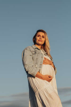 Portrait of Pregnant Young Caucasian Woman Holding Her Belly on the Background of Blue Skyの写真素材