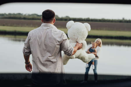 Young Father Holding Teddy Bear in Hand in Front of His Family, Dad Prepared a Present for His Little Daughter, View Through Car Windowの写真素材