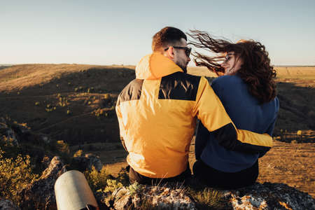 Back View of Traveler Couple Hugging and Sitting on the Edge of Hill During Sunset, Wind Blowing Womans Hairの写真素材