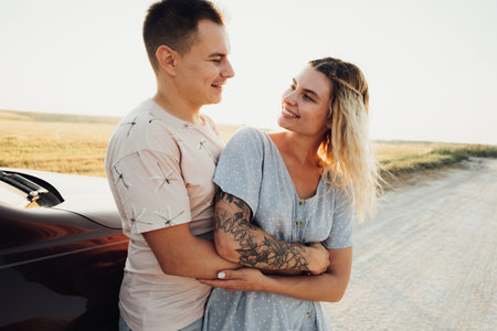 Cheerful Young Couple Hugging Near the Car, Tattooed Woman and Caucasian Man Enjoying Road Trip at Sunsetの写真素材