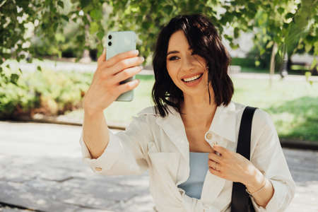 Cheerful Young Woman Making Video Call on Smartphone Outdoors Among Trees in the Park at Summer...