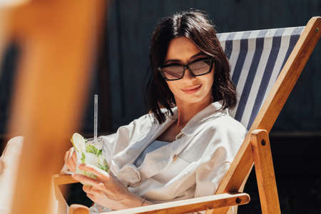 Portrait of Young Brunette Woman in Sunglasses Drinking Cocktail While Sitting on Terrace Outdoors...