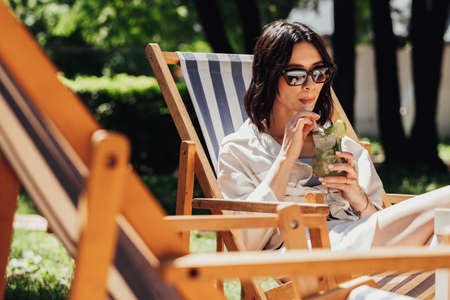 Young Brunette Woman in Sunglasses Drinking Cocktail While Sitting on Terrace Outdoors, Girl Enjoying Summer Sunny Dayの写真素材