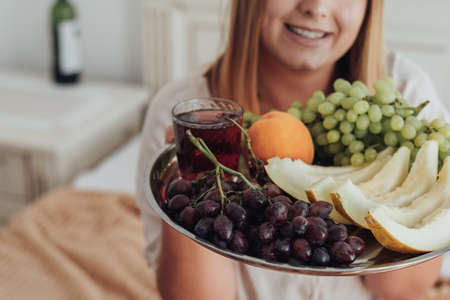 Unrecognisable Young Woman Holding Tray of Fruit in Hotel Room, Bottle of Wine Stands on the Backgroundの写真素材