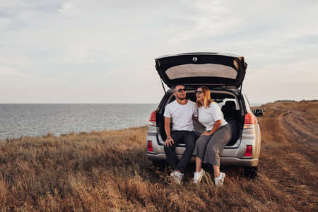 Happy Young Couple Sitting in Open Trunk of the SUV Car, Man and Woman Enjoying Their Road Trip...