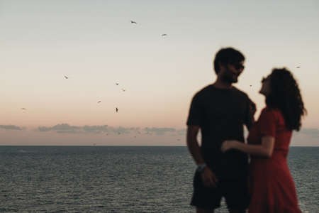 Unfocused Blurred Couple Standing on the Background of Sea After Sunset at Evening, Seagulls Flying Over Waterの写真素材