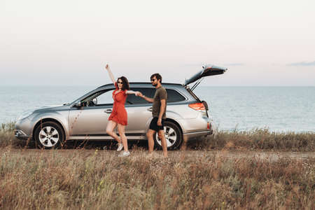 Youth Couple Having Fun on the Background of the Sea, Man and Woman Enjoying Road Trip Near Their SUV Car at Sunsetの写真素材