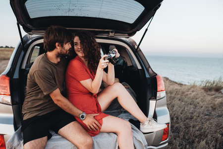Man and Woman Sitting Inside Opened Trunk of Their SUV Car, Young Couple Enjoying Road Trip Along Sea at Sunsetの写真素材
