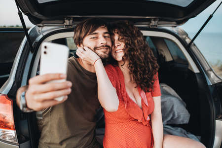 Happy Caucasian Couple Man and Curly Brunette Woman Making Selfie on Smartphone While Sitting in Trunk of the SUV Carの写真素材