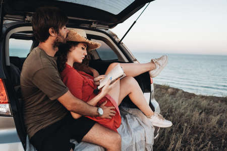 Man and Woman Sitting Inside Opened Trunk of Their SUV Car, Young Couple Enjoying Road Trip Along Sea at Sunsetの写真素材