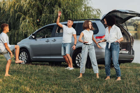 Happy Four Members Family Having Fun Time Outdoors, Mother Dancing with Teenage Daughter While Father Playing with Son, Minivan Car on the Backgroundの写真素材