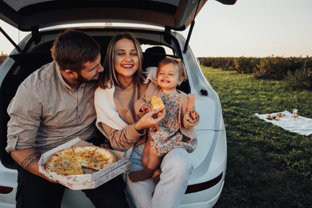 Happy Young Family Sitting in the Trunk of SUV Car and Eating Pizza, Mom Dad and Baby Daughter...
