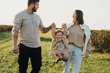Happy Young Family Outdoors, Mother and Father Lifting Their Baby Daughter by Hands at Sunset in the Fieldの写真素材