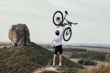 Professional Male Cyclist in Helmet Raising Hand with Bicycle while Standing on the Top of Hill, Giant Stone, Miracle of Nature on the Backgroundの写真素材