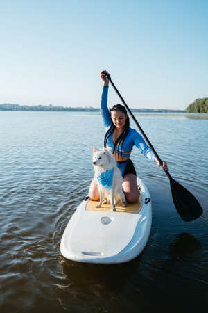 Young Woman with Dreads Paddleboarding with Her Pet on the City Lake, Snow-White Japanese Spitz Dog...