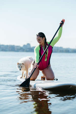 Happy Cheerful Woman Paddleboarding on the City Lake at Early Morning with Her Dog Japanese Spitz Sitting on the Sup Boardの写真素材