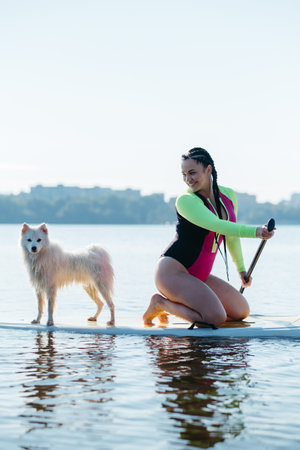 Happy Cheerful Woman Paddleboarding on the City Lake at Early Morning with Her Dog Japanese Spitz Sitting on the Sup Boardの写真素材