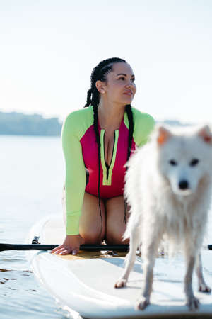 Portrait of Young Woman with Dreadlocks in Swimwear Sitting on the Sup Board with Her Dog Japanese Spitzの写真素材