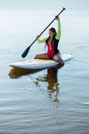 Young Woman with Dreadlocks in Swimwear Sitting on the Sup Board, Female Paddleboarding on the Lake at Sunriseの写真素材