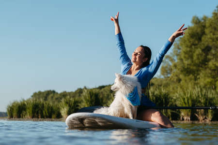 Happy Young Woman Raising Hands Up While Sitting on the Sup Board with Her Dog Japanese Spitz